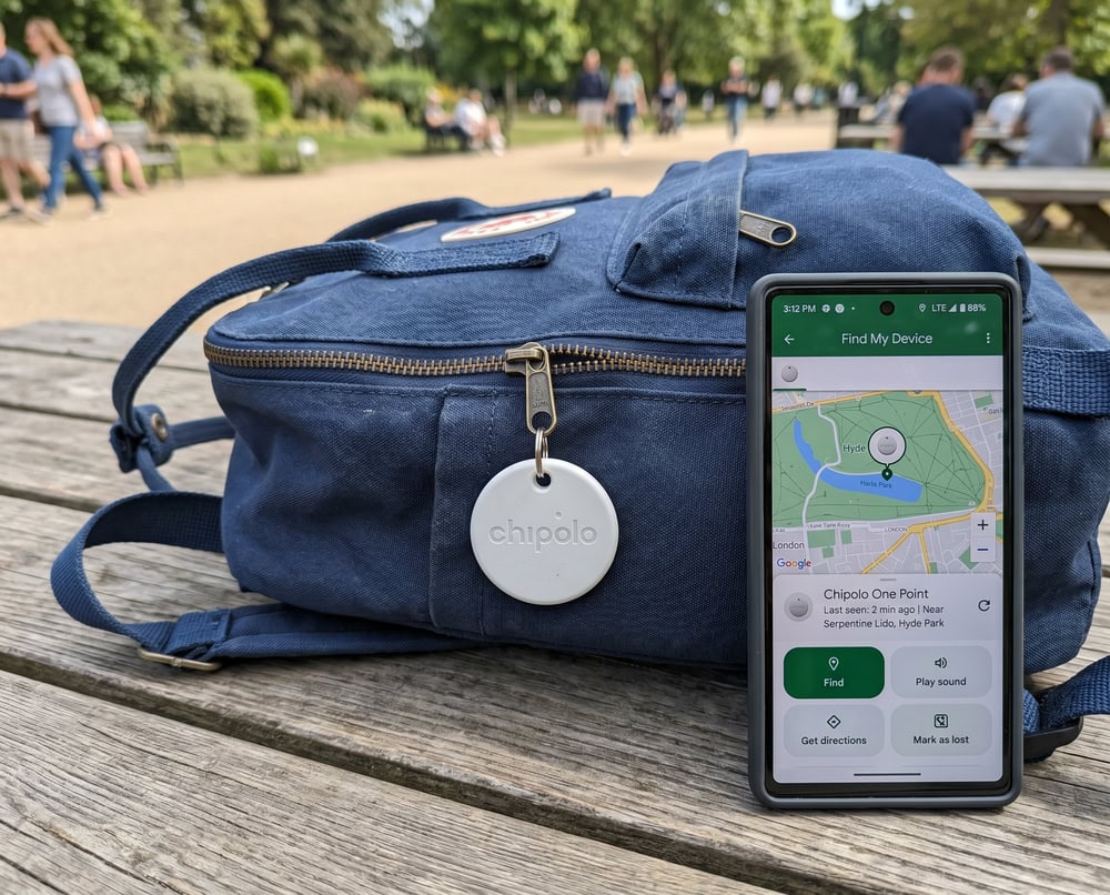 A white Chipolo One Point tag on a backpack zipper beside an Android phone showing its last-seen location in the Google Find Hub app.