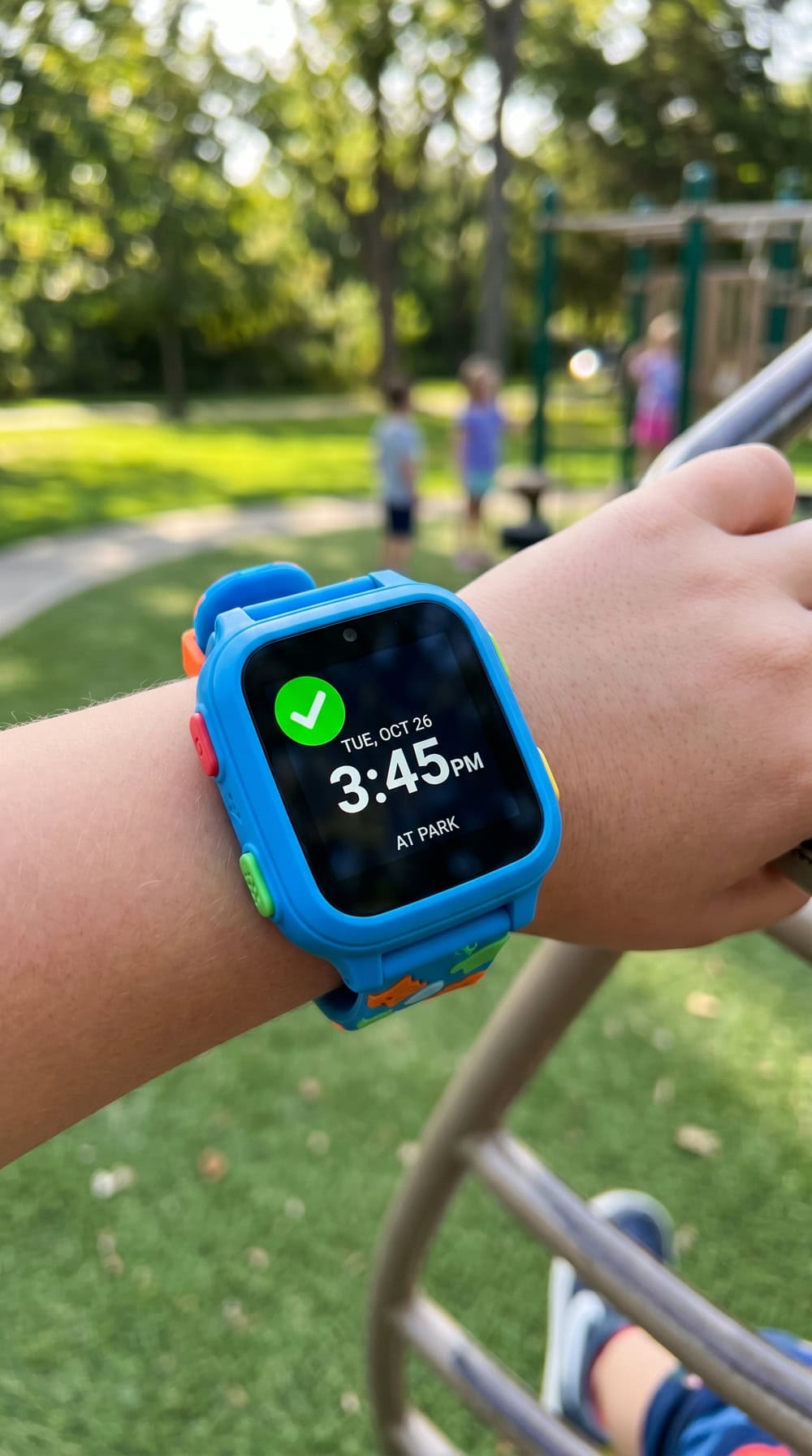 A child's wrist wearing a TickTalk 5 GPS watch showing the time and a green safe-zone status icon, photographed at a park.