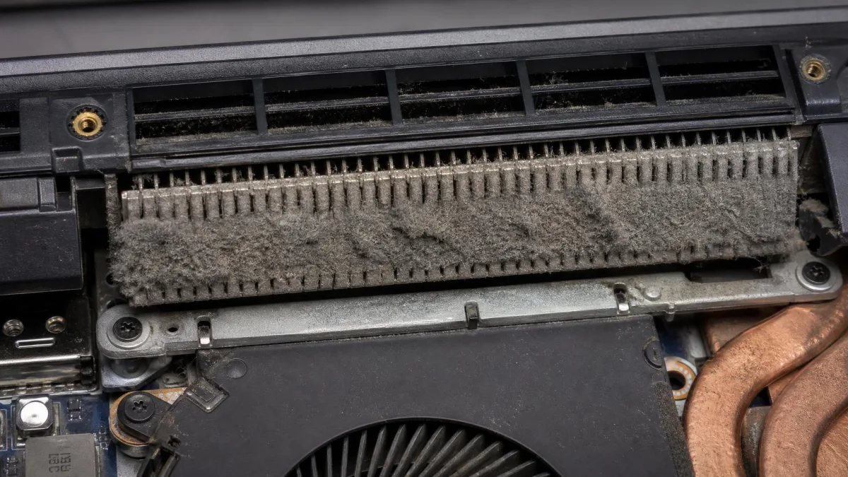 Close-up of a laptop's exhaust vent with a thick mat of grey dust across the heatsink fins, photographed with the bottom panel removed.