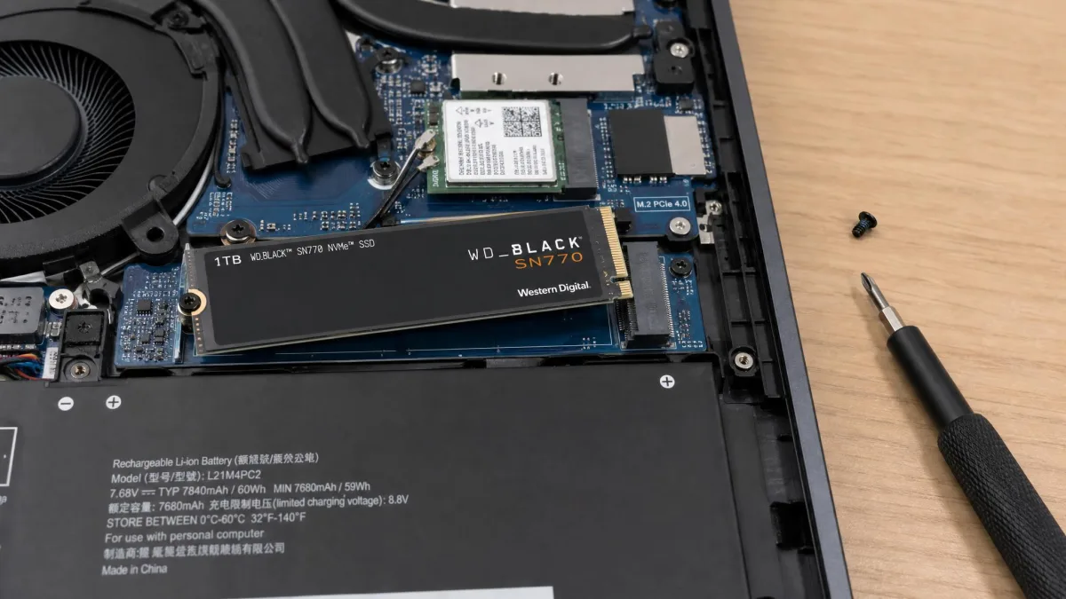 An open laptop underside with an M.2 SSD being seated at an angle into its slot, the retaining screw and a small screwdriver visible beside it.