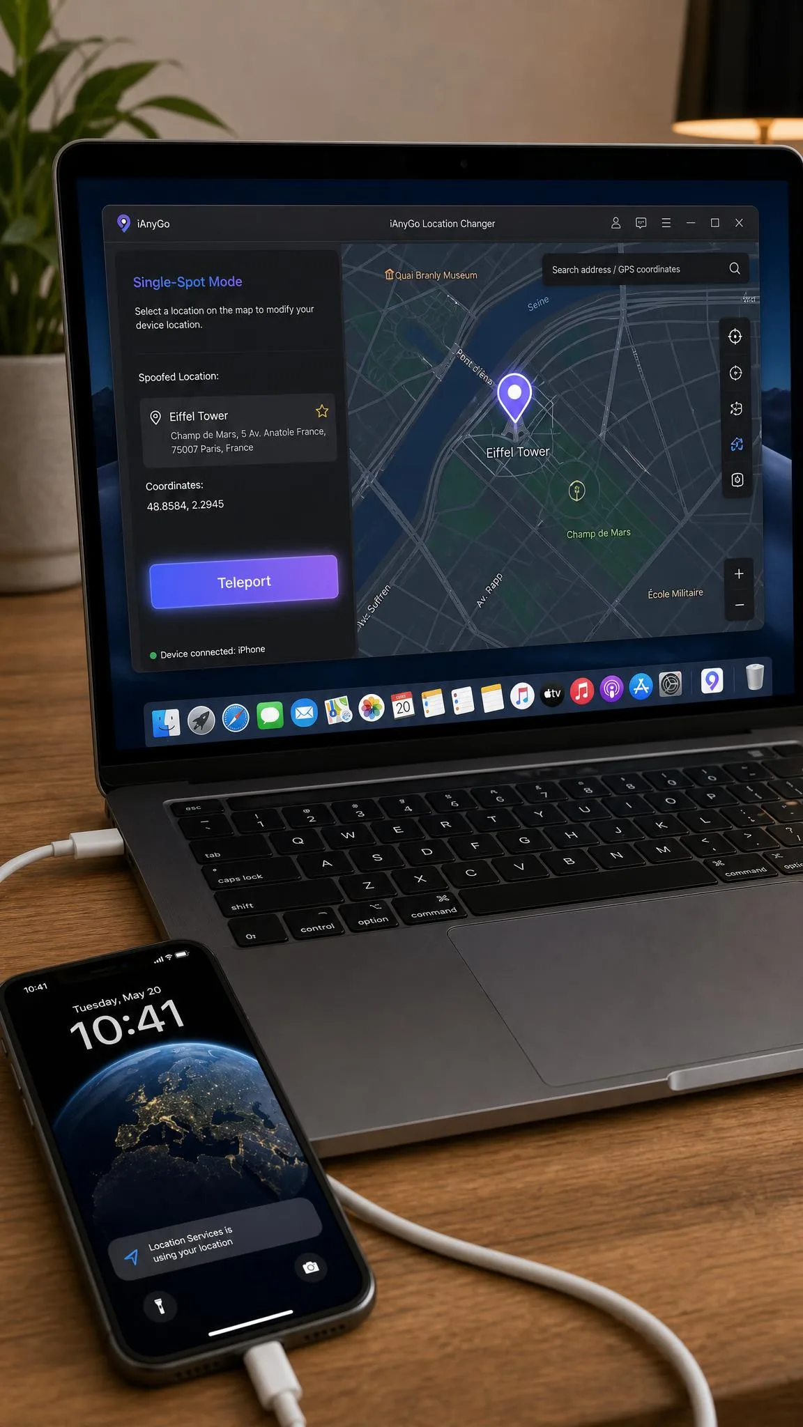 iPhone connected to a laptop with a USB cable while a location changer desktop app shows a spoofed pin on a city map with the teleport button highlighted.