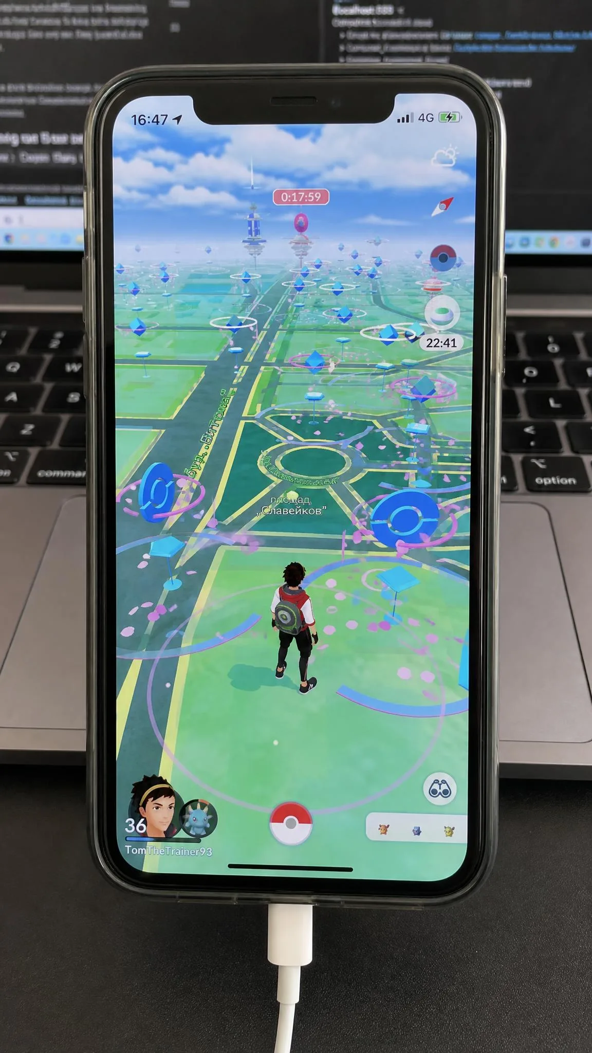 Pokemon GO map screen on an iPhone showing an avatar standing in a foreign city centre surrounded by PokeStops, with the phone plugged into a laptop.