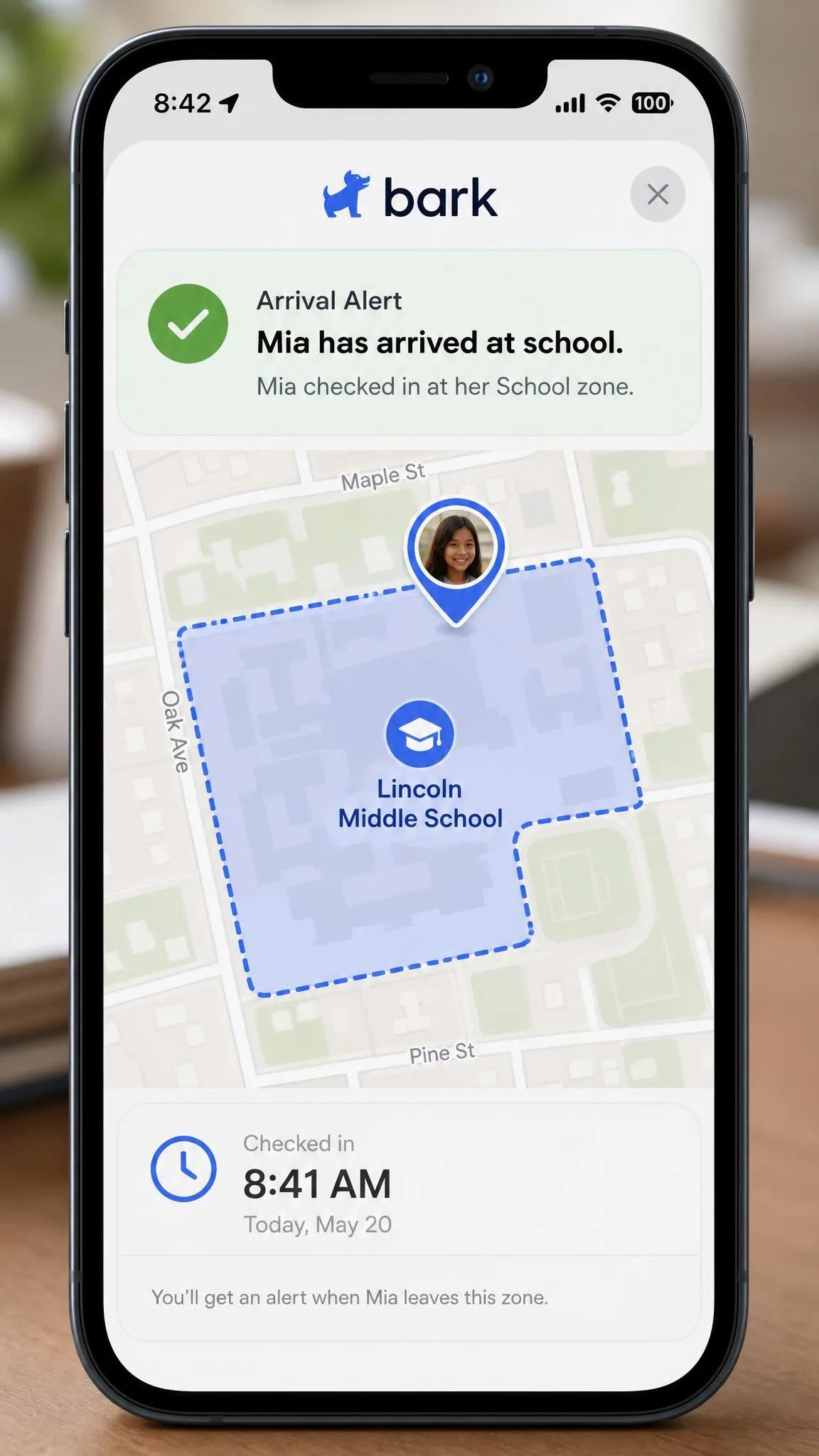 A phone showing a Bark arrival alert on a map, with a geofenced school zone outlined and a check-in time listed beneath the notification.