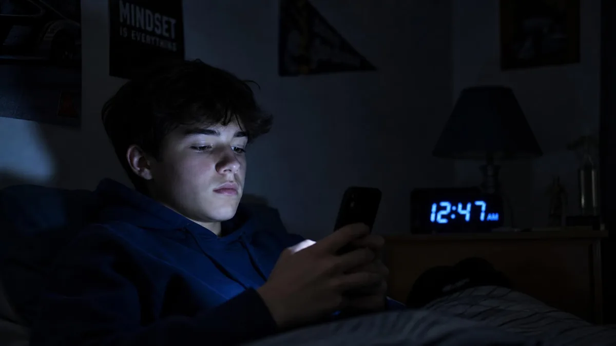 Teenage boy alone in his bedroom at night, face lit by his phone, a clock showing a late hour.