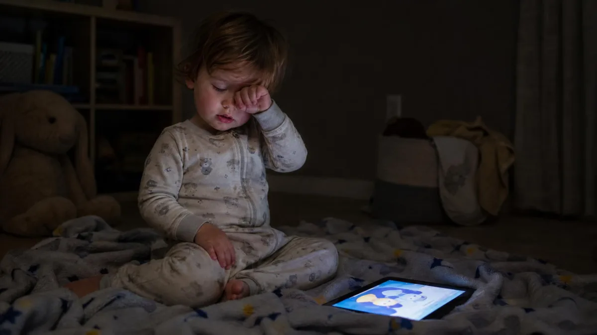 A sleepy toddler rubbing an eye in a dim room with a glowing tablet on the blanket beside them.